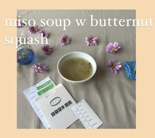 A bowl of miso soup with butternut squash, plated on a linen tablecloth surrounded by purple daisies.