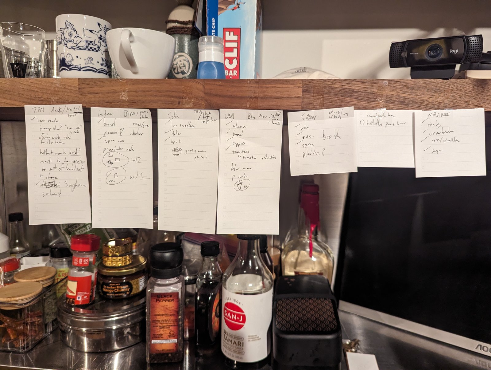 Index cards taped up under a kitchen shelf, one per country, with handwritten ingredient and dish notes for the dinner.
