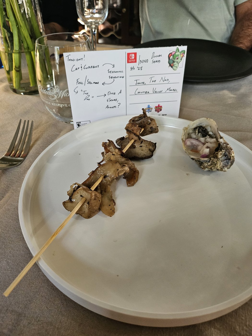 Grilled mushroom skewers and a single dressed oyster, plated beside the postcard menu.