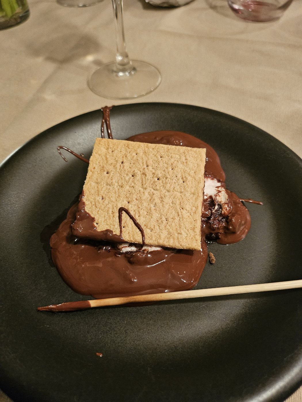 A s'more plated on a dark dish: graham cracker over a pool of melted chocolate and jam, with a roasting skewer.