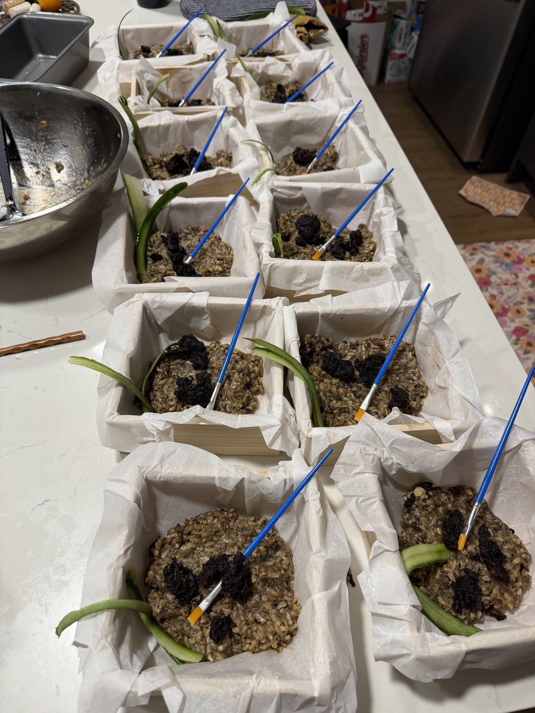 A row of paper-lined crates, each holding a portion of chicken ballotine over mushroom soil with a scallion and a paintbrush staged like an excavation site.