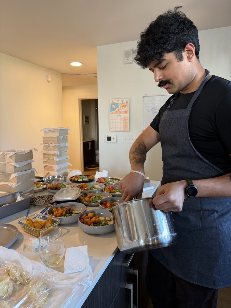 A cook in an apron plating dishes at the kitchen counter, surrounded by stacked takeout boxes and small bowls of ingredients.